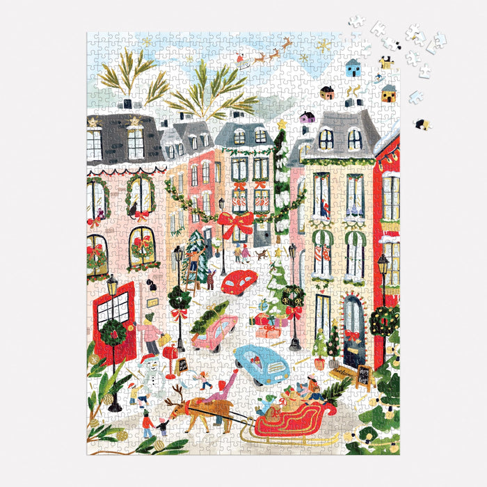 The Merriest Village 1000 Piece Foil Puzzle by Sabina Fenn features a festive town square with decorated buildings, wreaths, ice skaters, cars, and snowy streets. Some pieces are missing in the upper right corner.