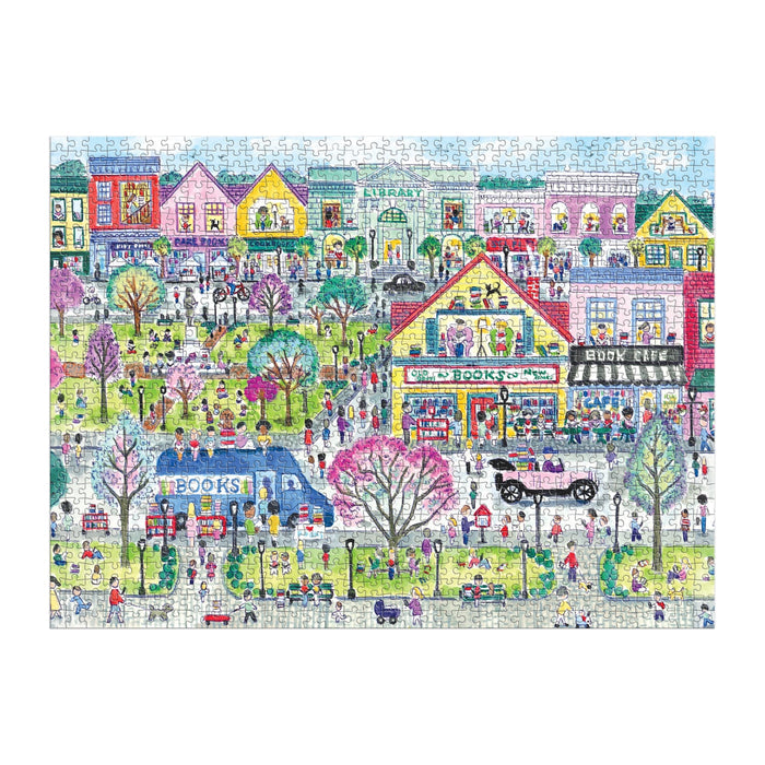 The Michael Storrings Book Town 1000 Piece Puzzle by Michael Storrings features a lively town scene with people, a bookshop, café, bookmobile, vintage car, trees, and vibrant buildings—perfect for book lovers celebrating community.