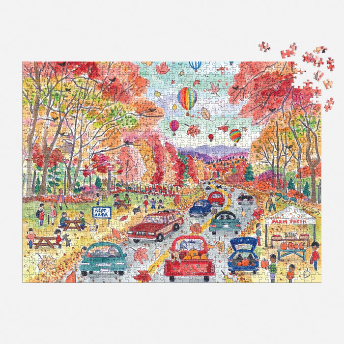 The Michael Storrings Leaf Peeping 1000 Piece Puzzle by Michael Storrings features cars on a tree-lined autumn road, people picnicking and buying food, and hot air balloons drifting overhead—a vibrant fall scene to assemble.