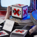 A Mini Poorly Explained Movies - Horror Edition game box by Brass Monkey rests on film reels, with playing cards showing vague horror movie clues—ideal for trivia or parties—all against a blue-lit backdrop.