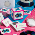 A box labeled Mini Poorly Explained Movies – Y2K Edition by Brass Monkey sits among clue cards, a computer mouse, CDs, and retro tech on a pink and blue background—ideal for your next pop culture trivia game night.