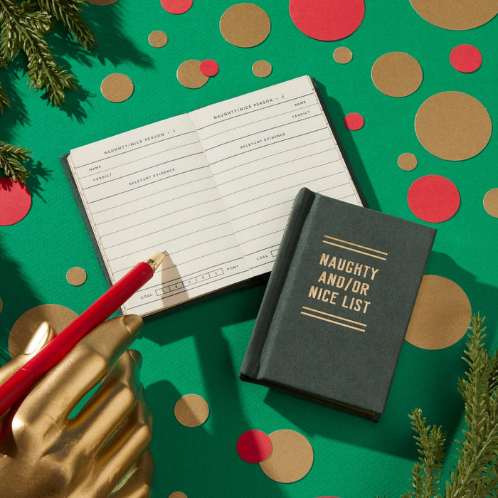 A gold mannequin hand holds a red pencil over an open Naughty and/or Nice Tiny Diary by Brass Monkey, set on a green background with festive accents. A closed pocket-sized version of the diary lies nearby, both featuring the playful title.