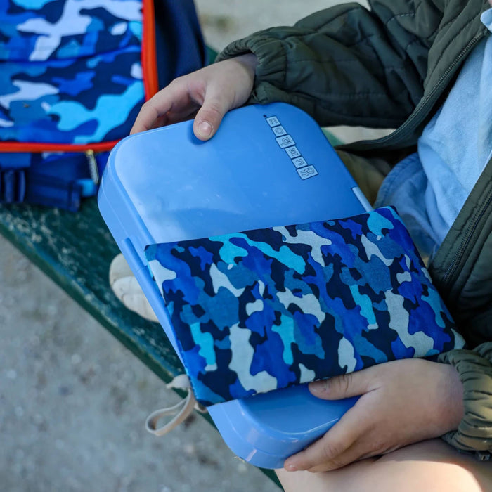 Blue plastic bento lunch box with a blue camouflage-patterned gel ice pack. held by a child sitting cross-legged on a bench.