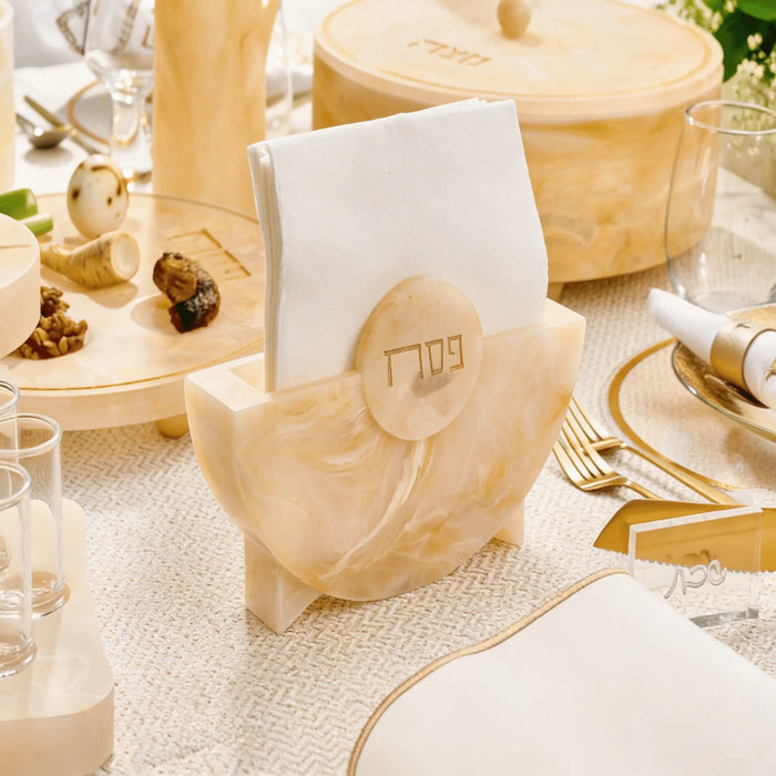 Beige resin Pesach napkin holder with embossed design on white table setting with gold accents and place cards