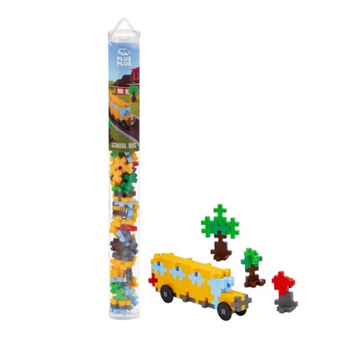 A clear plastic tube of small building blocks stands next to a assembled yellow school bus model with small trees, on a plain white background. The tube label shows the brand name 'Plus Plus'.