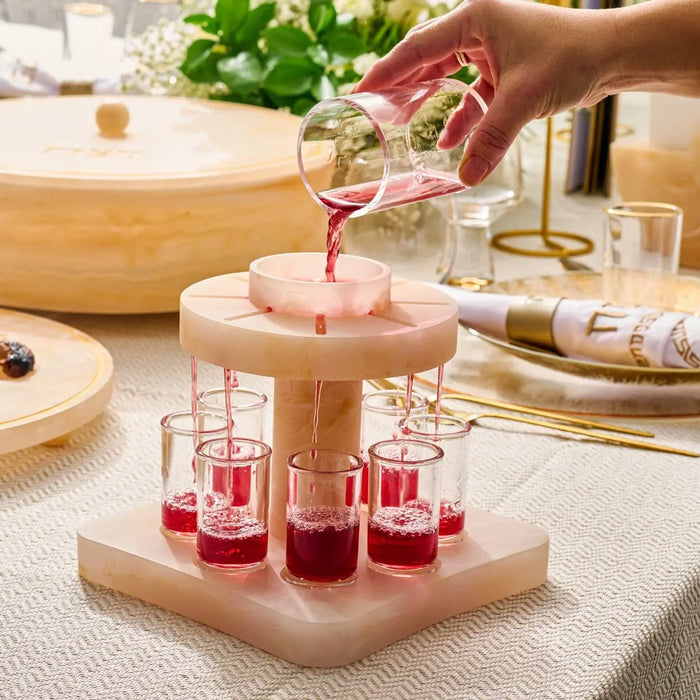Wooden resin Kiddush fountain with pink glass cups arranged on tiered wooden base, displayed on kitchen counter with Shabbat items.