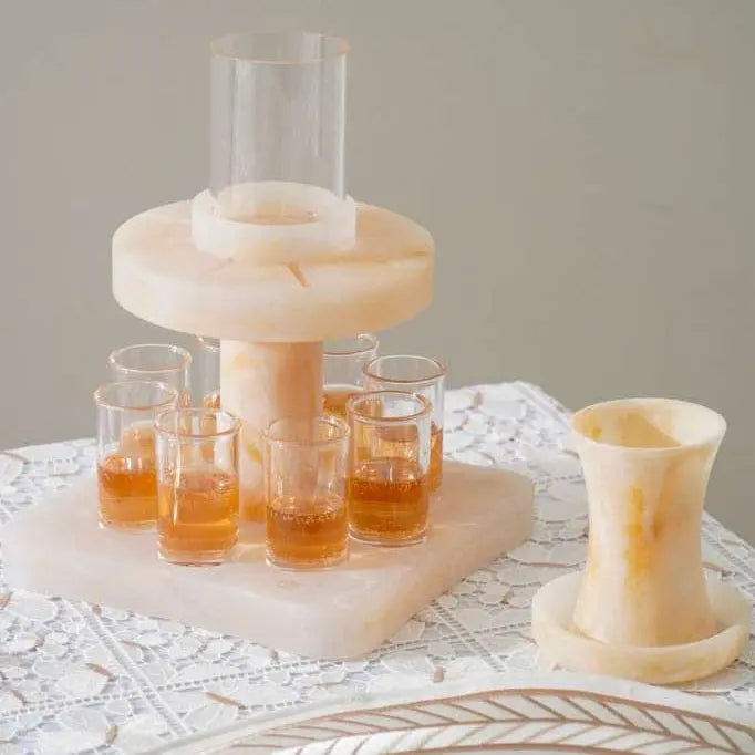 Beige resin Kiddush fountain with multiple cup holders and matching serving cups, displayed on white decorative surface for Shabbat wine service.