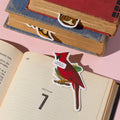 The Salty Birds Magnetic Bookmark Set by Brass Monkey features a red cardinal-shaped bookmark with a “What is this stuff?” speech bubble, sitting in an open book as bird-shaped bookmarks peek from two nearby closed books on a pale pink surface.