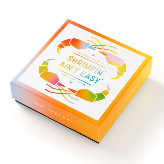 The Shrimpin Aint Easy Origami Kit by Brass Monkey comes in a colorful square box with a geometric shrimp design, An Origami Kit text, and playful orange-to-yellow packaging for fun shrimp origami inspiration.