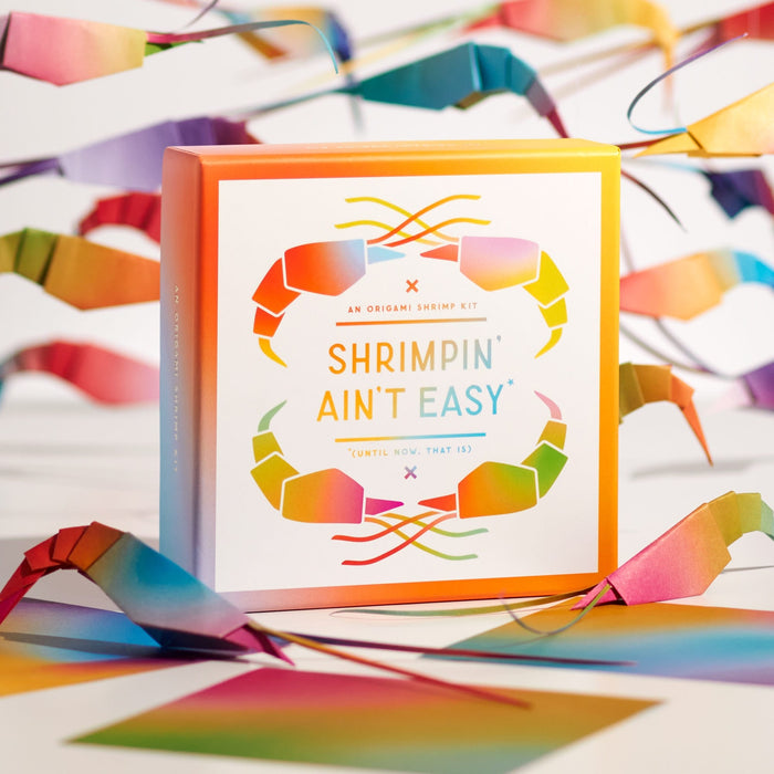 A colorful box labeled Shrimpin Aint Easy by Brass Monkey is surrounded by rainbow shrimp illustrations, matching paper shrimp decorations, and the Shrimpin Aint Easy Origami Kit on a white background.