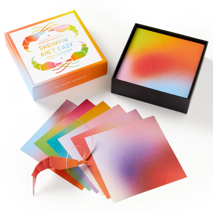 The Shrimpin Aint Easy Origami Kit by Brass Monkey features a decorative box, gradient papers displayed, and a completed shrimp with googly eyes placed next to the open kit.