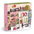 The Snowfall on Main Street 1000 Piece Foil Puzzle by Nysha Lilly showcases a festive, snowy main street with colorful houses, children playing, and shimmering foil accents highlighting holiday décor and the sparkling Christmas tree.