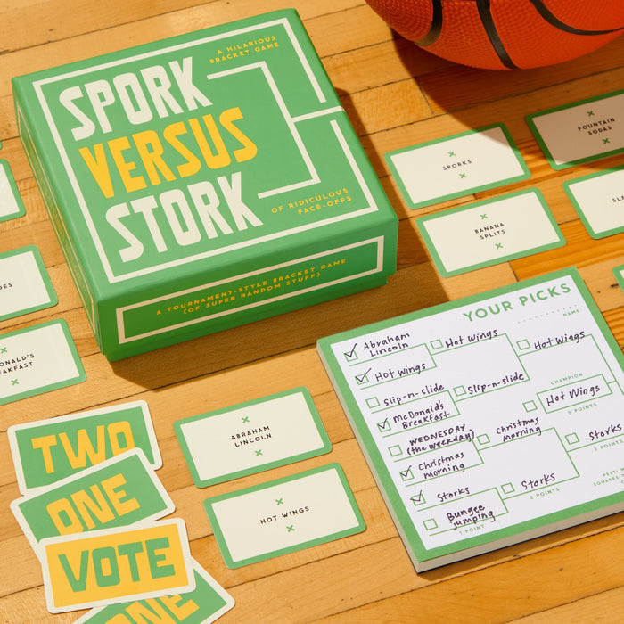 Brass Monkey’s Spork Versus Stork Social Game is displayed on a wooden surface with cards, score sheet, and basketball. Players write picks for fun categories like Hot Wings or Abraham Lincoln, then reveal choices as each card is played.