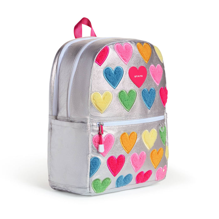A metallic silver backpack decorated with colorful felt hearts in pink, blue, green, yellow, and orange. The backpack features a top zipper, a front zippered pocket with a pink zipper pull, and a pink top handle, front angle view shown against a plain white background.