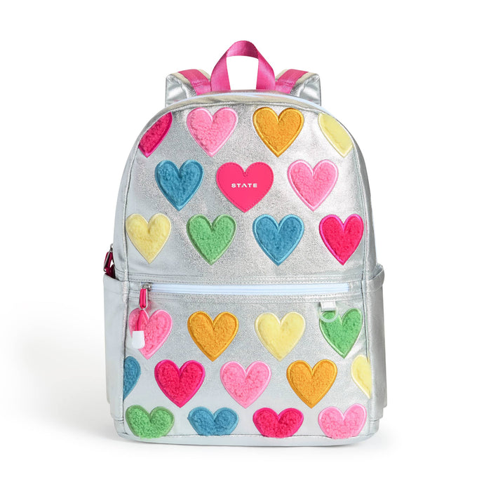 A metallic silver backpack decorated with colorful felt hearts in pink, blue, green, yellow, and orange. The backpack features a top zipper, a front zippered pocket with a pink zipper pull, and a pink top handle, front view shown against a plain white background.