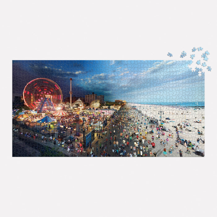 The Stephen Wilkes Day To Night Coney Island 1500 Piece Puzzle by Stephen Wilkes features a vivid beach sunset with a lit-up carnival and Ferris wheel; a few pieces are missing in the top right corner.
