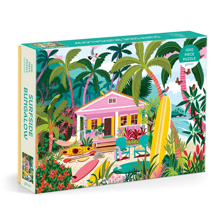 The Julia Cornelius Surfside Bungalow 1000 Piece Puzzle features a tropical beach scene with a pink bungalow, palm trees, surfboards, kayaks, and beach chairs by the ocean under a bright blue sky.