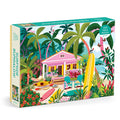 The Julia Cornelius Surfside Bungalow 1000 Piece Puzzle features a tropical beach scene with a pink bungalow, palm trees, surfboards, kayaks, and beach chairs by the ocean under a bright blue sky.