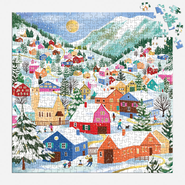 The Swiss Alpine Village 500 Piece Foil Puzzle by Joanna Clay features a vibrant winter scene with snowy houses, trees, playful villagers, and a shining sun. The mostly finished puzzle has a few missing pieces in the top right corner.