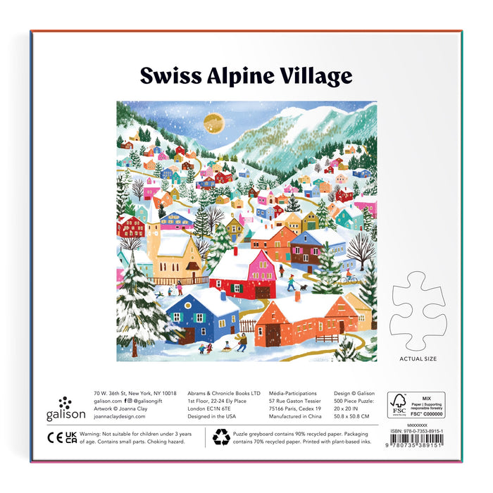 Experience winter charm with Joanna Clay's Swiss Alpine Village 500 Piece Foil Puzzle, featuring a snowy mountain village, bright houses, pine trees, and people enjoying winter fun beneath a starry sky. Includes a sample puzzle piece for size reference.