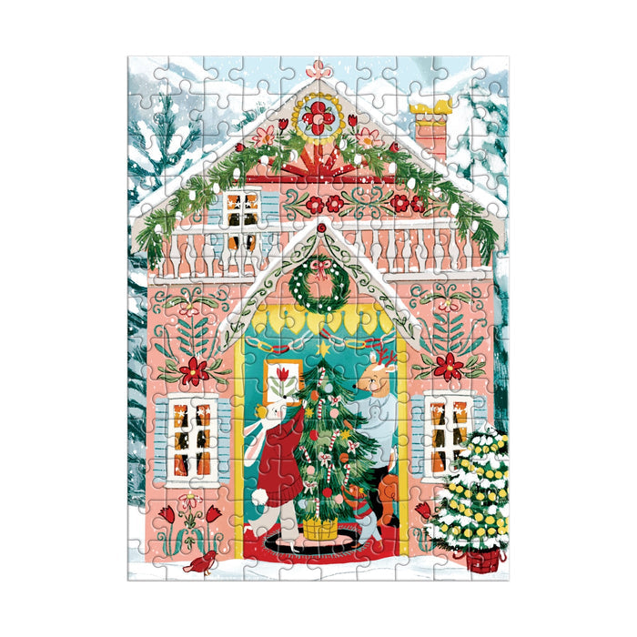 The Swiss Chalet 130 Piece Ornament Puzzle by Joanna Clay features a festive pink house adorned with garlands, wreaths, and lights, a family decorating a Christmas tree inside, snow on the roof and ground, and a lit tree outside.