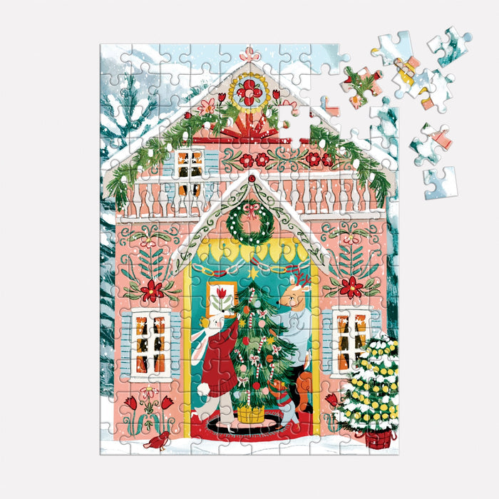 The Joanna Clay Swiss Chalet 130 Piece Ornament Puzzle features a festive house with a decorated Christmas tree, two people hanging ornaments, snowy trees and bushes outside, and several unfinished pieces in the top right corner.