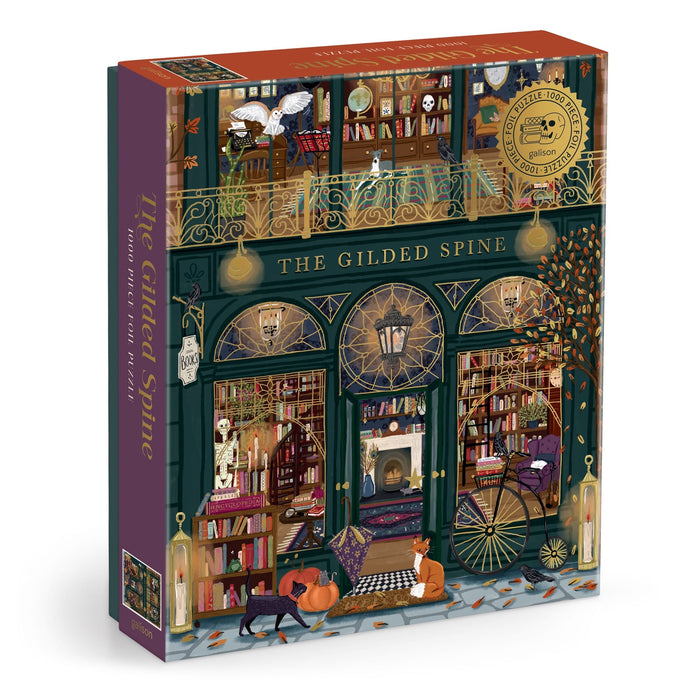 The Gilded Spine 1000 Piece Foil Puzzle by Sally Wilson depicts a charming bookstore with large windows, shelves of books, autumn leaves, cozy lights, a black cat, and a dog inside and outside the shop.