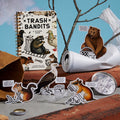 Brass Monkey's Trash Bandits Sticker Book sits with puffy stickers, a fake tree branch, bottle cap, crumpled cup, and coin. Inside, raccoon and bear stickers have funny speech bubbles for a playful vibe.