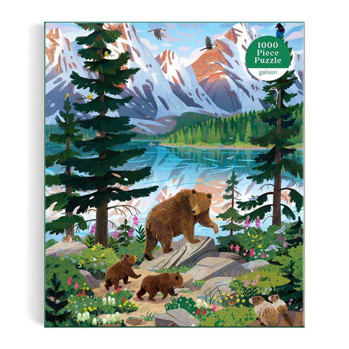The Valley of the Peaks 1000 Piece Puzzle by Kim Smith features a brown bear with three cubs in a vibrant alpine forest by a lake, set against snow-capped mountains and birds in the background.