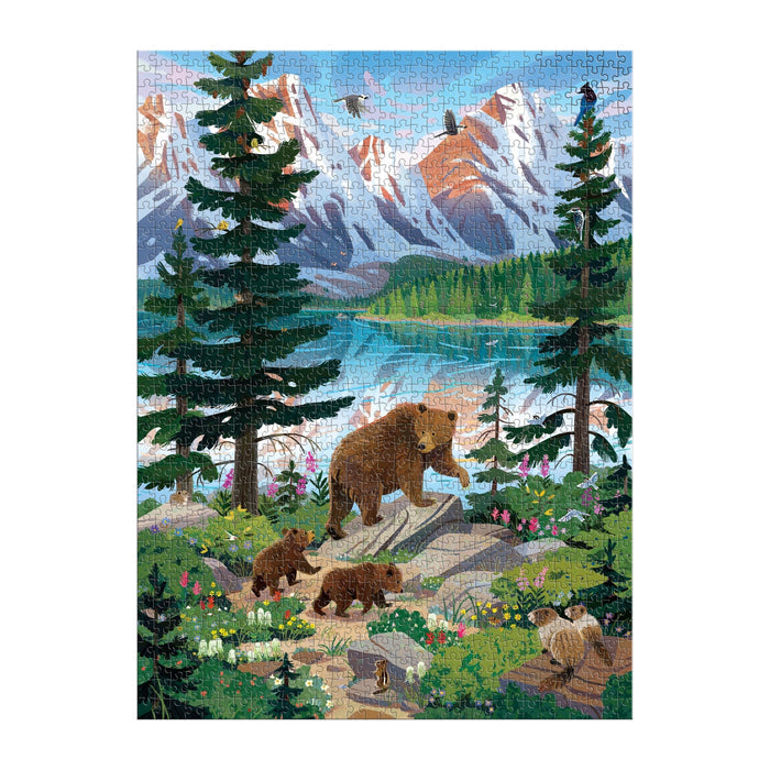 The Valley of the Peaks 1000 Piece Puzzle by Kim Smith features a bear with three cubs on a forest trail by a lake, with snowy mountain peaks, wildflowers, green trees, and a clear blue sky creating a beautiful nature scene.