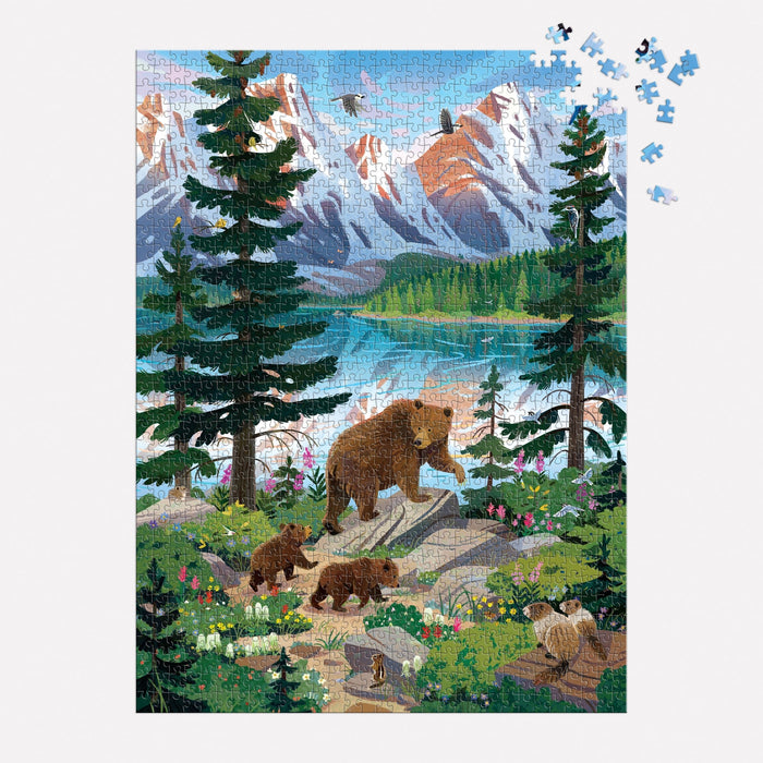 The Valley of the Peaks 1000 Piece Puzzle by Kim Smith features a scenic mountain landscape with a lake, pine trees, and a bear family on a rocky path. A few pieces remain to be placed in the top right corner.