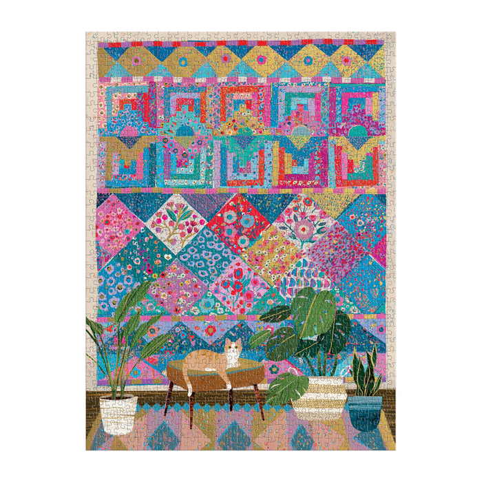 The Victoria Ball Quilters Corner 1000 Piece Foil Puzzle by Victoria Ball features a vibrant quilt tapestry with intricate floral patterns above a wooden table, while a tan and white cat lounges below among green potted plants on a wooden floor.