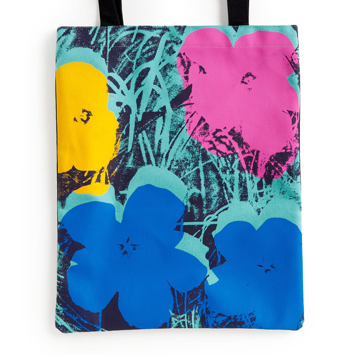 The Warhol Flowers Volume 3 Canvas Tote Bag by Andy Warhol Foundation For The Visual Arts features a pop art design with large yellow, pink, and blue flowers on a green and black background with abstract grass-like patterns and black handles.
