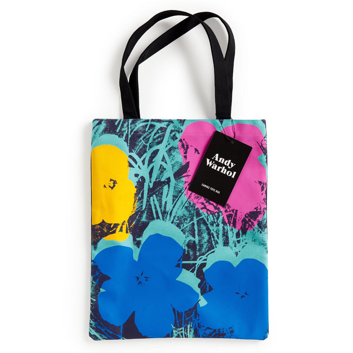 The Warhol Flowers Volume 3 Canvas Tote Bag by the Andy Warhol Foundation features blue, yellow, and pink flowers on green, with black handles and an attached Andy Warhol tag.