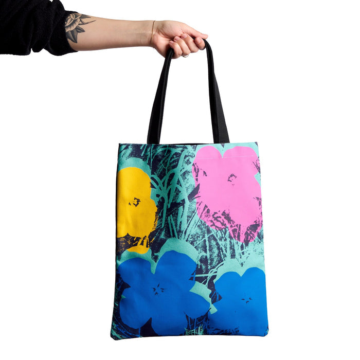 A hand holds the Warhol Flowers Volume 3 Canvas Tote Bag by Andy Warhol Foundation For The Visual Arts, featuring vibrant yellow, pink, and blue floral art over a green and black background. The person's forearm is tattooed.