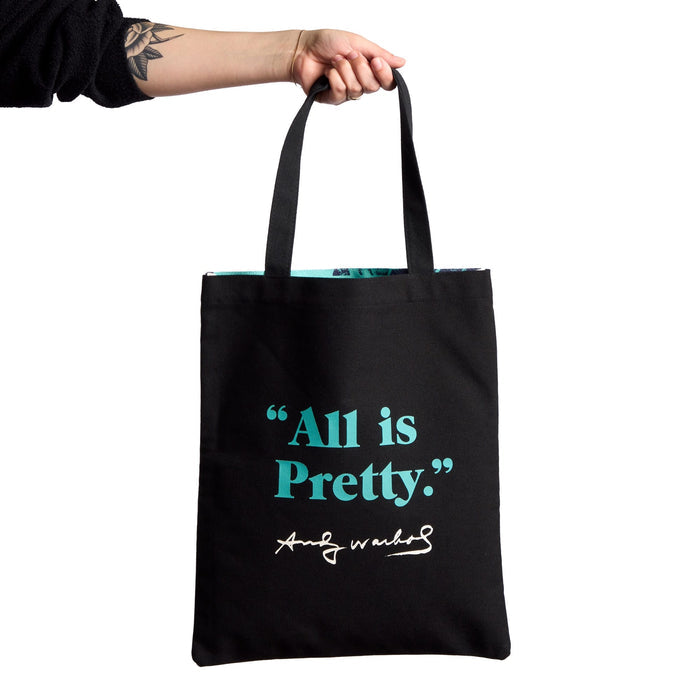 A hand holds the Warhol Flowers Volume 3 Canvas Tote Bag by Andy Warhol Foundation For The Visual Arts, featuring turquoise "All is Pretty." text and a white Andy Warhol signature on black canvas.