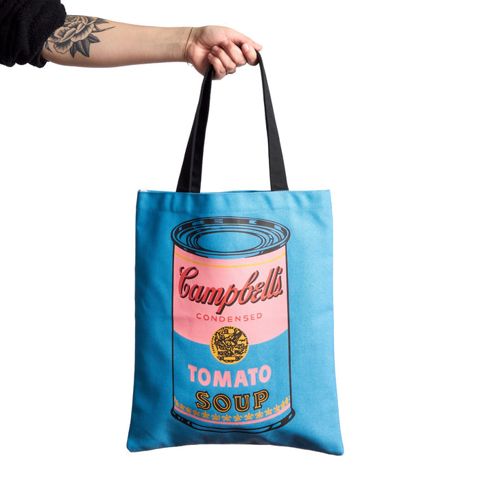 A hand with a floral tattoo holds the Warhol Soup Can Volume 3 Canvas Tote Bag by Andy Warhol Foundation For The Visual Arts, featuring a blue pop art Campbell’s tomato soup can design and black handles.