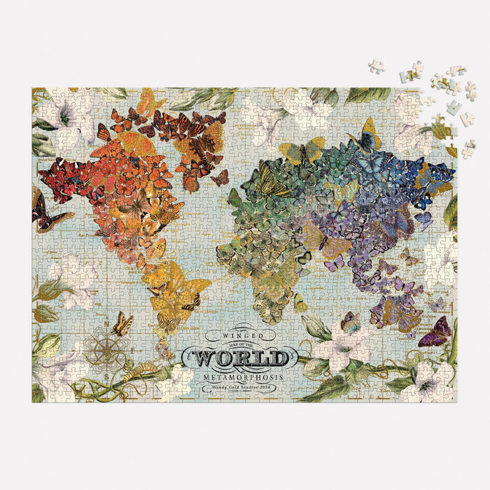 The completed Wendy Gold Winged Metamorphosis 1000 Piece Foil Puzzle by Wendy Gold features a world map made of vibrant butterfly wings, arranged by continent and ringed with flowers. Loose pieces appear in the upper right, with “Winged World Metamorphosis” below.